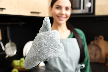 Woman in apron and oven glove showing thumb up indoors, focus on hand. Space for textの写真素材