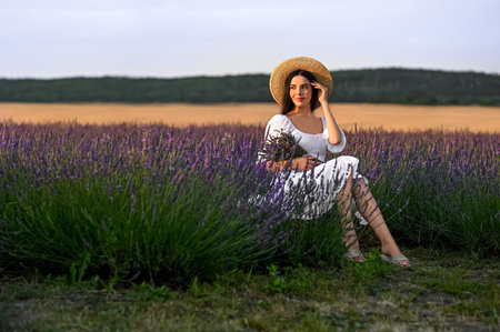 Beautiful young woman with bouquet sitting in lavender field at sunsetの写真素材