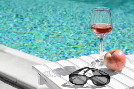 Glass of tasty rose wine, sunglasses and apple on wooden table near swimming pool, space for text