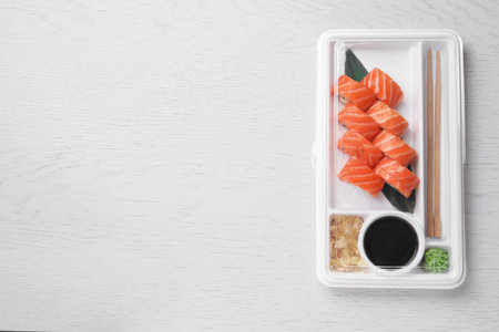 food delivery. Plastic container with delicious sushi rolls, soy sauce, ginger, wasabi, chopsticks on white wooden table, top view and space for textの写真素材
