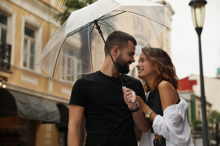 Young couple with umbrella enjoying time together under rain on city streetの写真素材