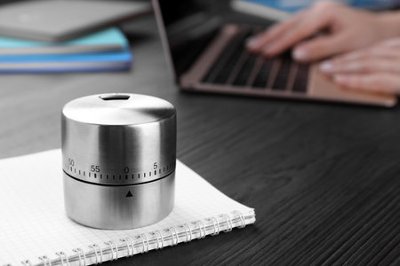 Woman working on laptop at wooden table, focus on kitchen timer. Space for text