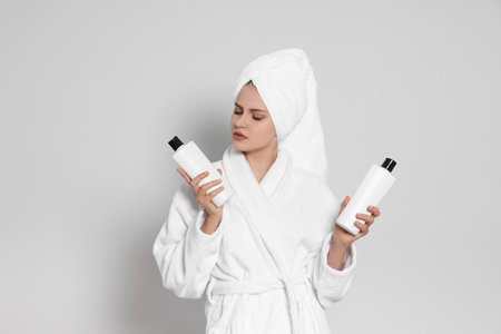 Beautiful young woman in bathrobe holding bottles of shampoo on light gray backgroundの写真素材