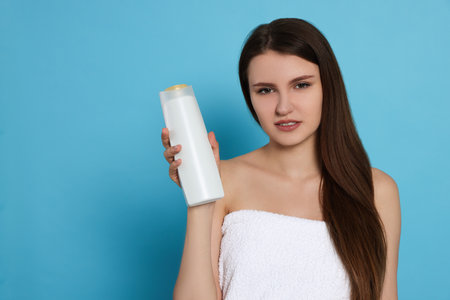 Beautiful young woman wrapped in towel holding bottle of shampoo on light blue background. Space for textの写真素材