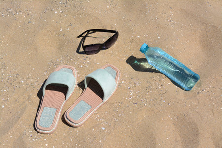 Stylish sunglasses, slippers and bottle of water on sand. beach accessoriesの写真素材