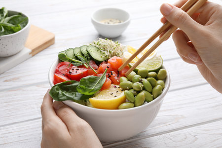 Woman eating delicious poke bowl with quail eggs, fish and edamame beans at white wooden table,...