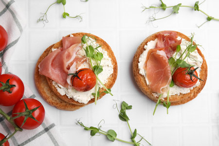 Tasty rusks with prosciutto, cream cheese and tomatoes on white table, flat layの写真素材
