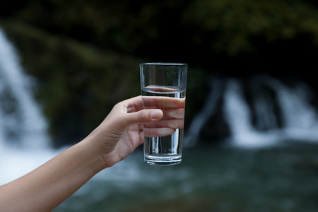 Woman holding glass of fresh water near waterfall, closeupの写真素材