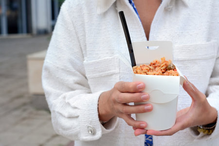Woman holding paper box of takeaway noodles with fork outdoors, closeup. street food