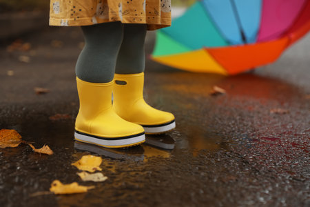 Little girl wearing yellow rubber boots standing in puddle outdoors, closeup
