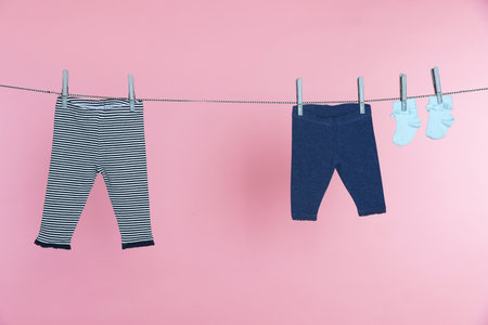 Different baby clothes drying on laundry line against pink background
