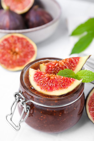 Glass jar of tasty sweet fig jam and fruits on white tiled table