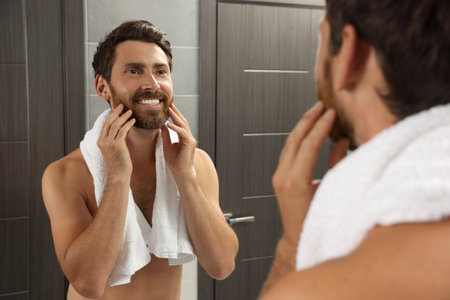 Handsome bearded man looking at mirror in bathroom near wooden doorsの写真素材