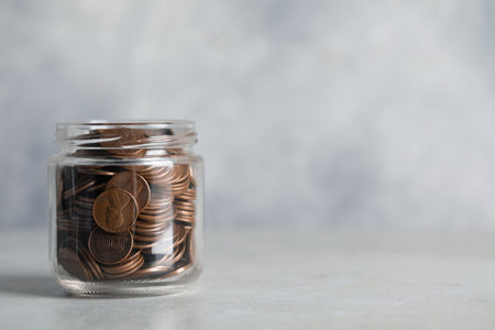 Glass jar with coins on light gray table, closeup. Space for text