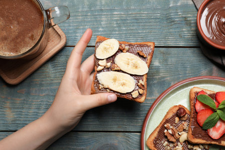 Woman holding tasty toast with chocolate spread, nuts and banana on wooden table, top view