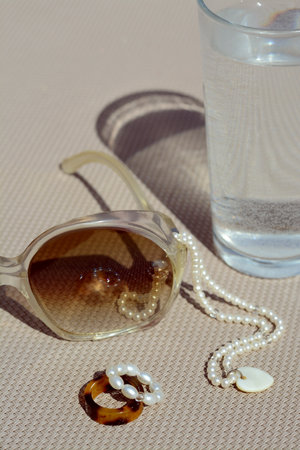 Stylish sunglasses, glass of water and jewelry on gray surface, closeup