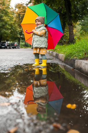 Cute little girl with colorful umbrella standing in puddle outdoorsの写真素材