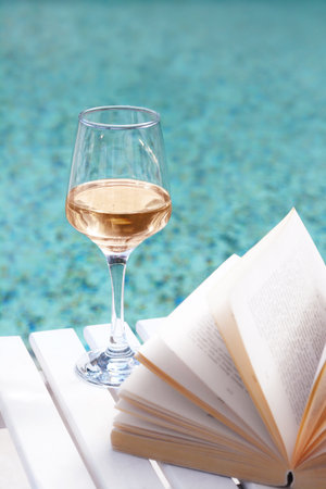 Glass of tasty wine and open book on wooden table near swimming poolの写真素材