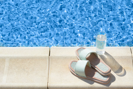 Stylish slippers and glass of water near outdoor swimming pool on sunny day, space for text
