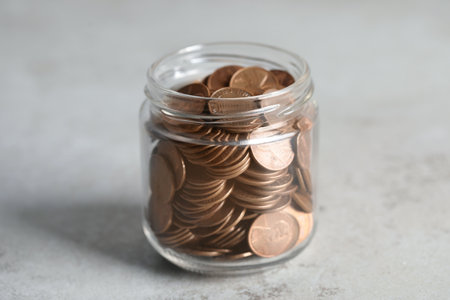 Glass jar with coins on light gray table, closeup