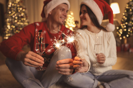 Couple in Santa hats holding sparkles and champagne glasses, focus on fireworks. christmas celebrationの写真素材