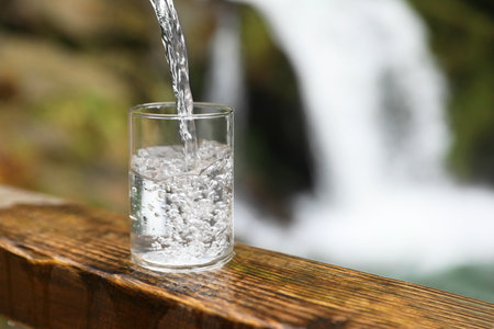 Fresh water pouring into glass on wooden surface near waterfall outdoors, closeup. Space for textの写真素材