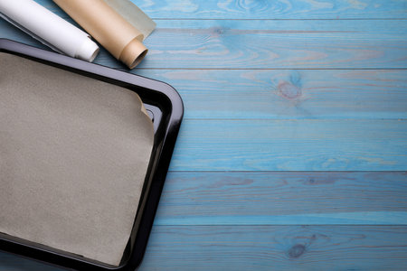 Rolls of parchment paper and baking pan on light blue wooden table, flat lay. Space for text