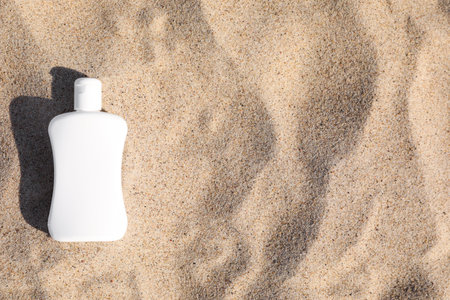 Blank white bottle of sunscreen on sand, top view. Space for text