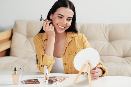 Beautiful young woman using eyelash curler while doing makeup at table indoorsの写真素材