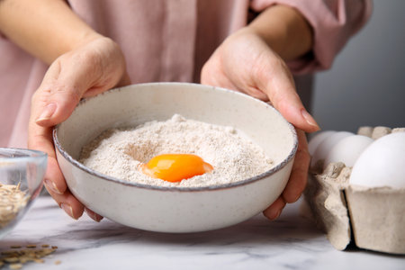 Woman holding ceramic bowl with flour and egg at white marble table in kitchen, closeup. Cooking oatmeal doughの写真素材