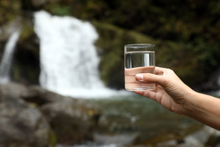 Woman holding glass of fresh water near waterfall outdoors, closeup. Space for textの写真素材
