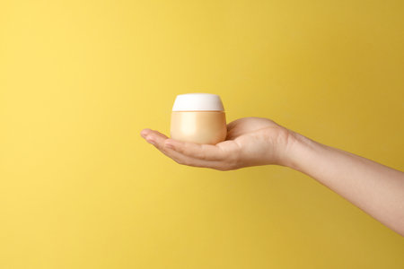 Woman holding jar of face cream on yellow background, closeup. Space for textの写真素材