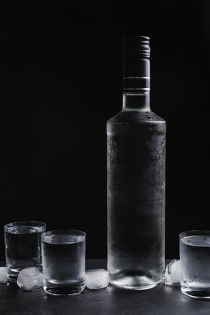 Bottle of vodka and shot glasses with ice on table against black backgroundの写真素材