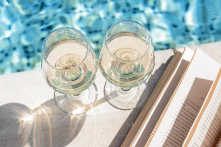 Glasses of tasty wine and open book on swimming pool edge, closeup