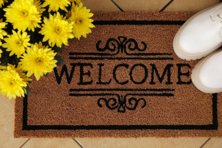 Door mat with word Welcome, stylish boots and beautiful flowers on floor, flat lay