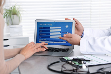 Doctor and patient using laptop to fill out medical card at table in clinic, closeup