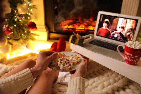 MYKOLAIV, UKRAINE - DECEMBER 23, 2020: Woman with cookies watching Harry Potter and Philosopher's Stone movie on laptop near fireplace at home, closeup. Cozy winter holidays atmosphereのeditorial素材