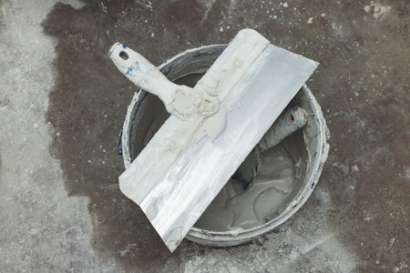 Bucket with plaster and putty knives on concrete floor, top view