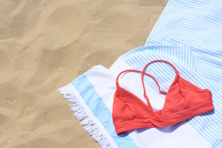 White and light blue striped towel with bikini top on sandy beach, flat lay. Space for text