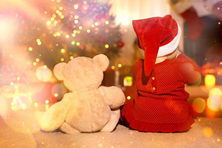 Baby wearing Santa hat and teddy bear in room decorated for Christmas, back view. Magical festive atmosphereの写真素材