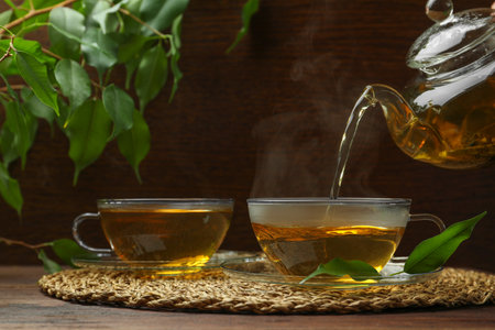 Pouring green tea into glass cup with saucer and leaves on wooden tableの写真素材
