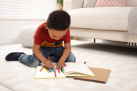 Cute African-American boy drawing in sketchbook with colorful markers on floor at home