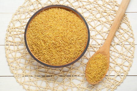 Bowl and spoon with uncooked bulgur on white wooden table, flat lay