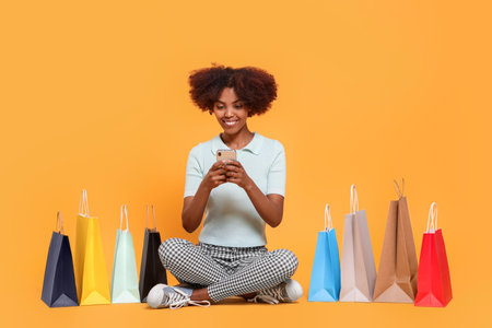 Happy African American woman with shopping bags and smartphone on orange backgroundの写真素材