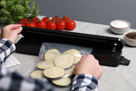 Woman packing cut eggplant into plastic bag using vacuum sealer on light gray marble table, closeupの写真素材
