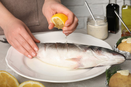 Woman squeezing lemon juice on fresh sea bass fish at light gray marble table, closeupの写真素材