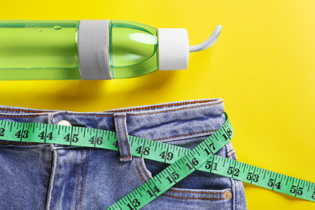 Jeans with measuring tape and bottle of water on yellow background, flat lay. weight loss conceptの写真素材
