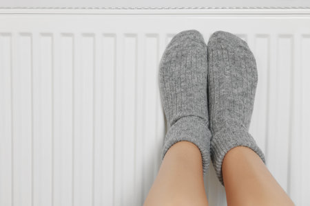 Woman warming legs on heating radiator near white wall, closeup. Space for textの写真素材