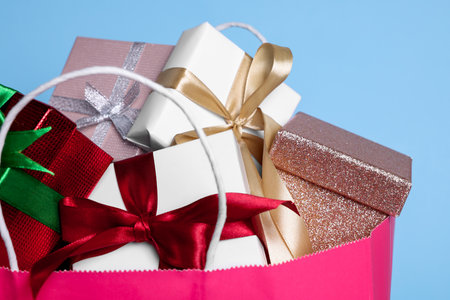 Pink paper shopping bag full of gift boxes on light blue background, closeup