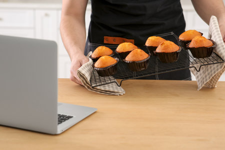 Man holding muffins near laptop at wooden table indoors, closeup. Time for hobby